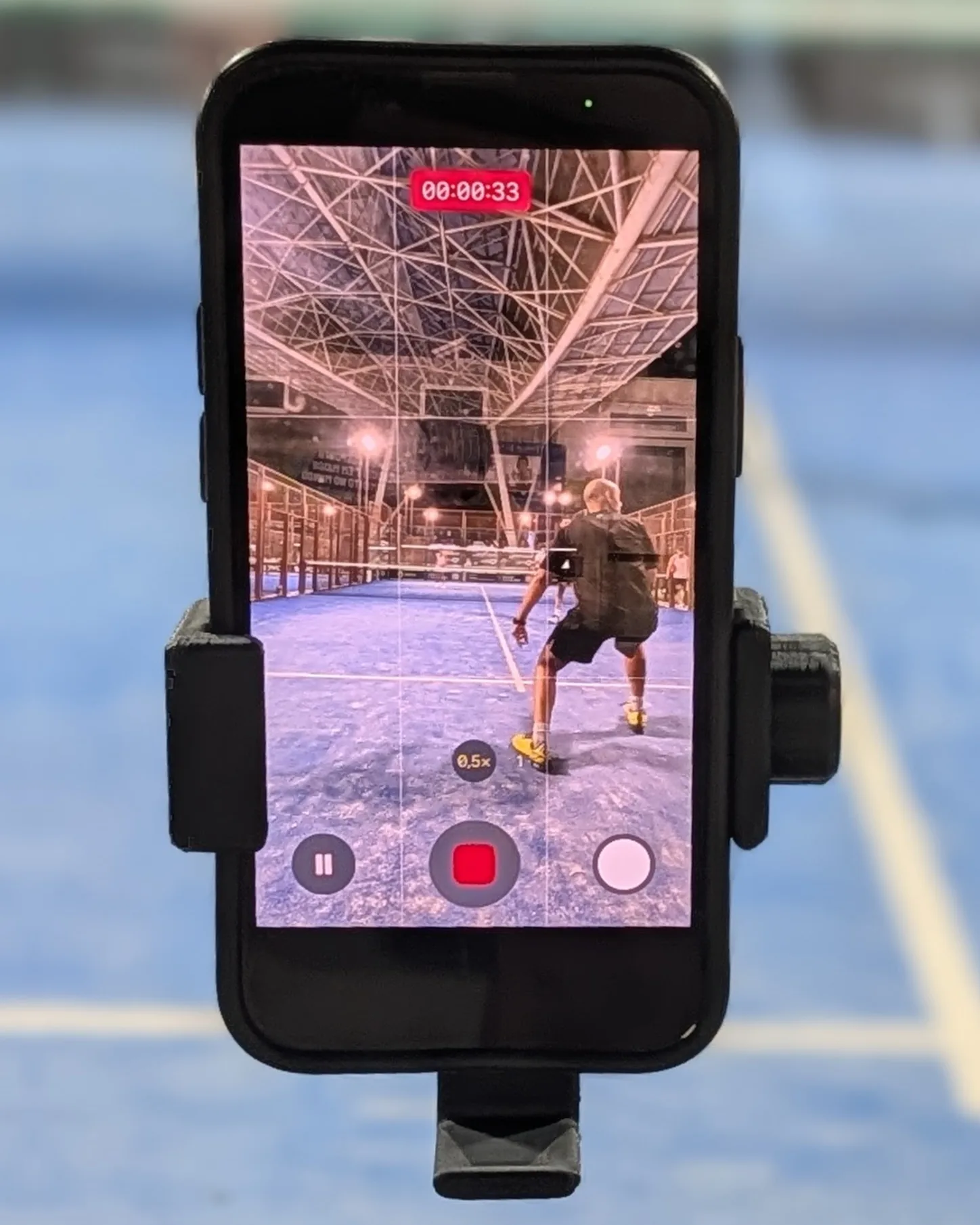 A phone in a Padel Holder on the glass at the back of an indoor padel court, recording the match behind it.